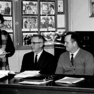 Five men sitting at desk with women standing, taking notes.Photo by Robert Mills