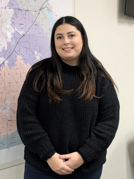 A photo of Kayla standing indoors against a wall with a map of Richland County in the background. She is smiling and wearing a black sweater with her hands clasped in front of her. 