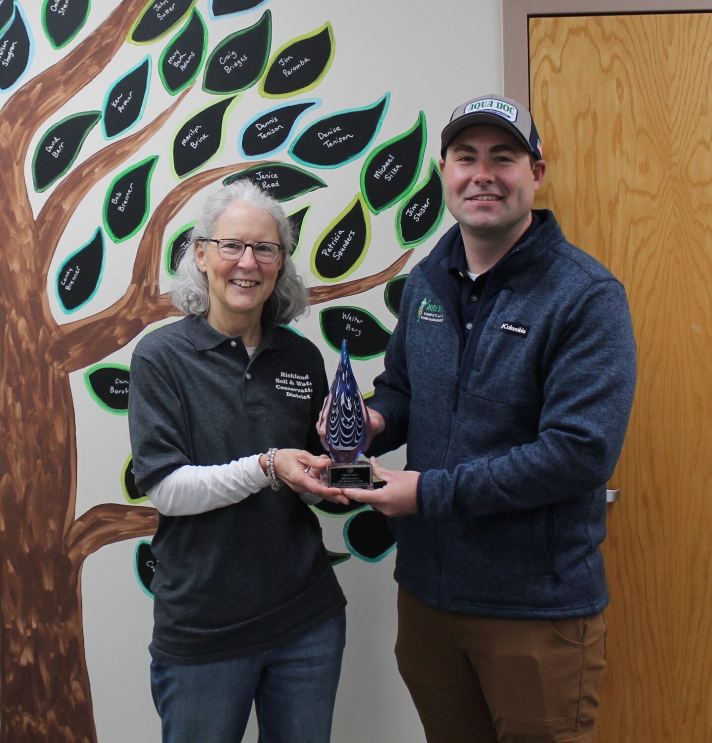Carter handing water droplet shaped award to Theresa. They're both smiling for the camera.