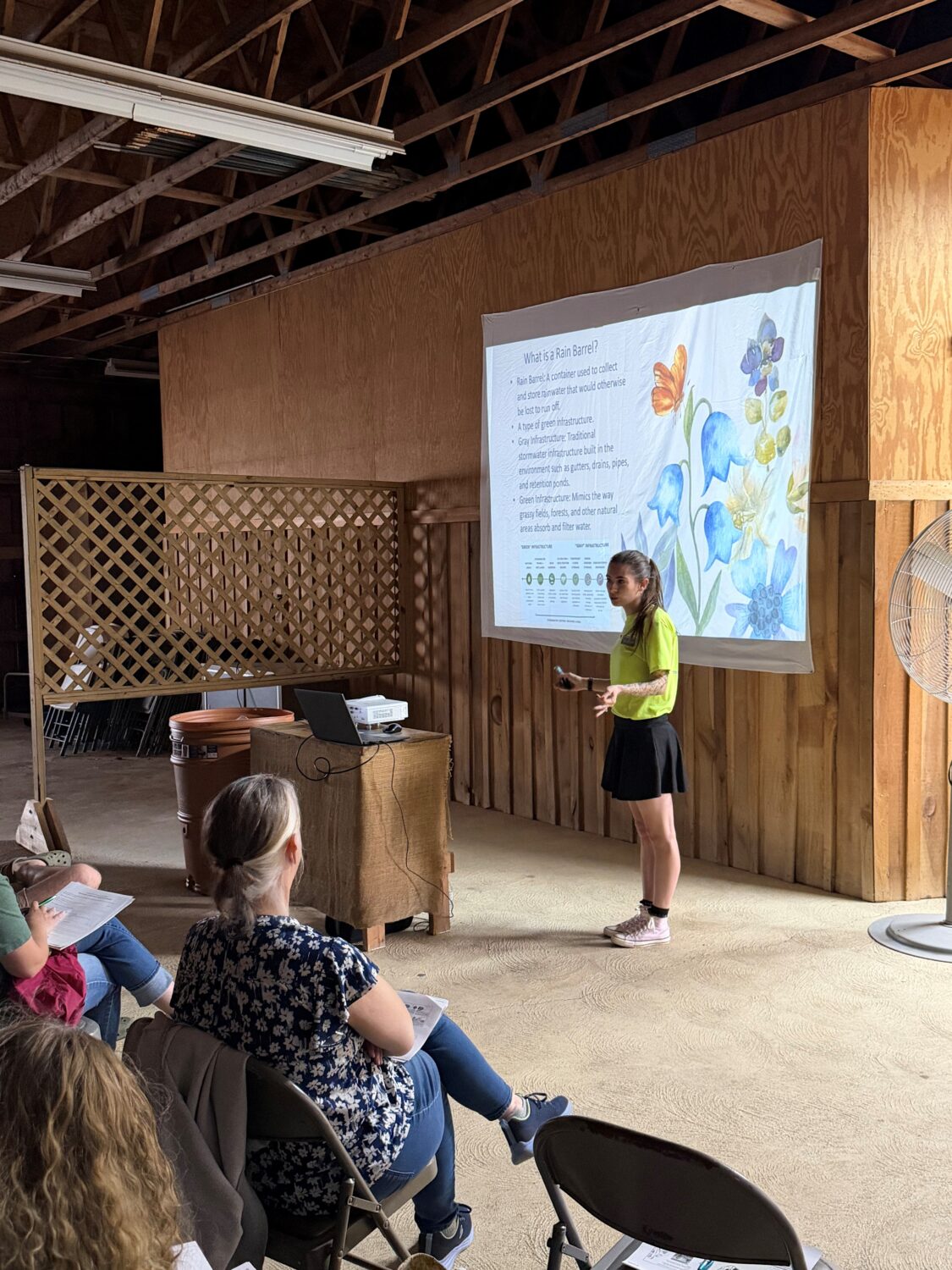 Zoe standing in front of a powepoint slide titled "what is a rain barrel". There are 2 audience members in the front row, 